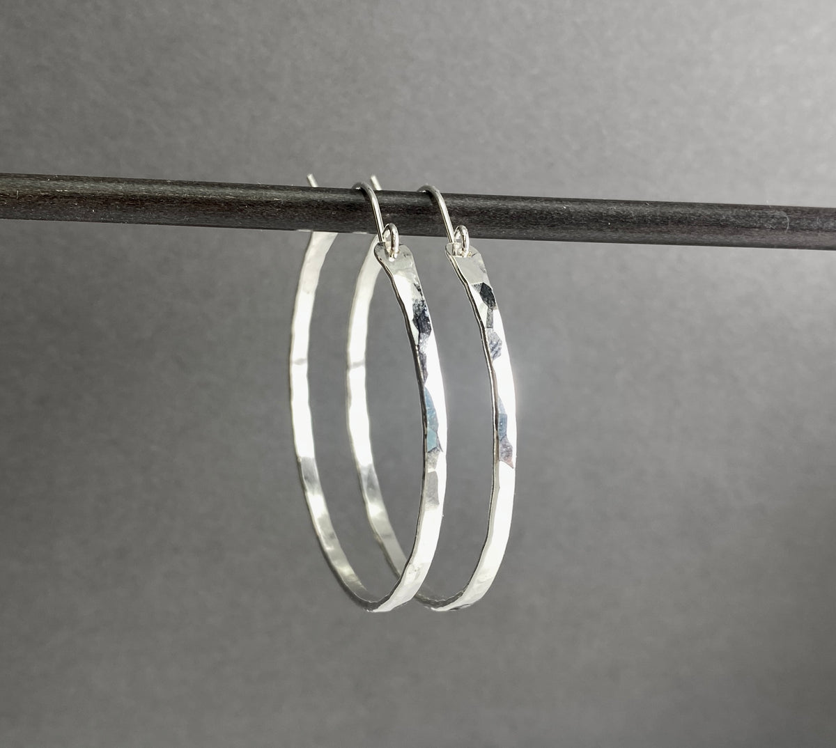 A pair of hammered silver hoop earrings hanging from a dark horizontal bar against a smooth gray background.