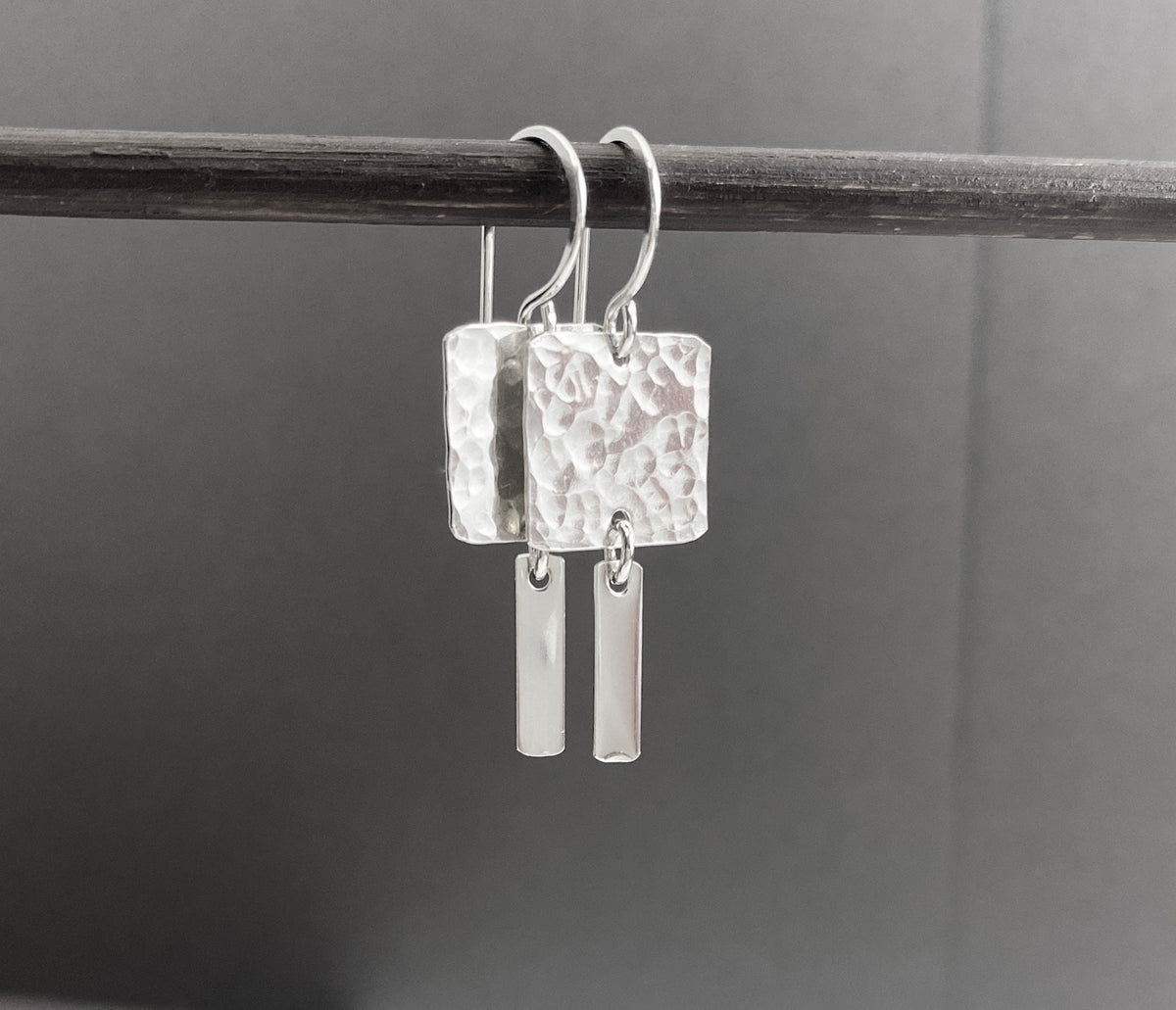 A pair of silver drop earrings with hammered square pendants and slim rectangular bar charms, hanging from a dark horizontal rod against a dark gray background.