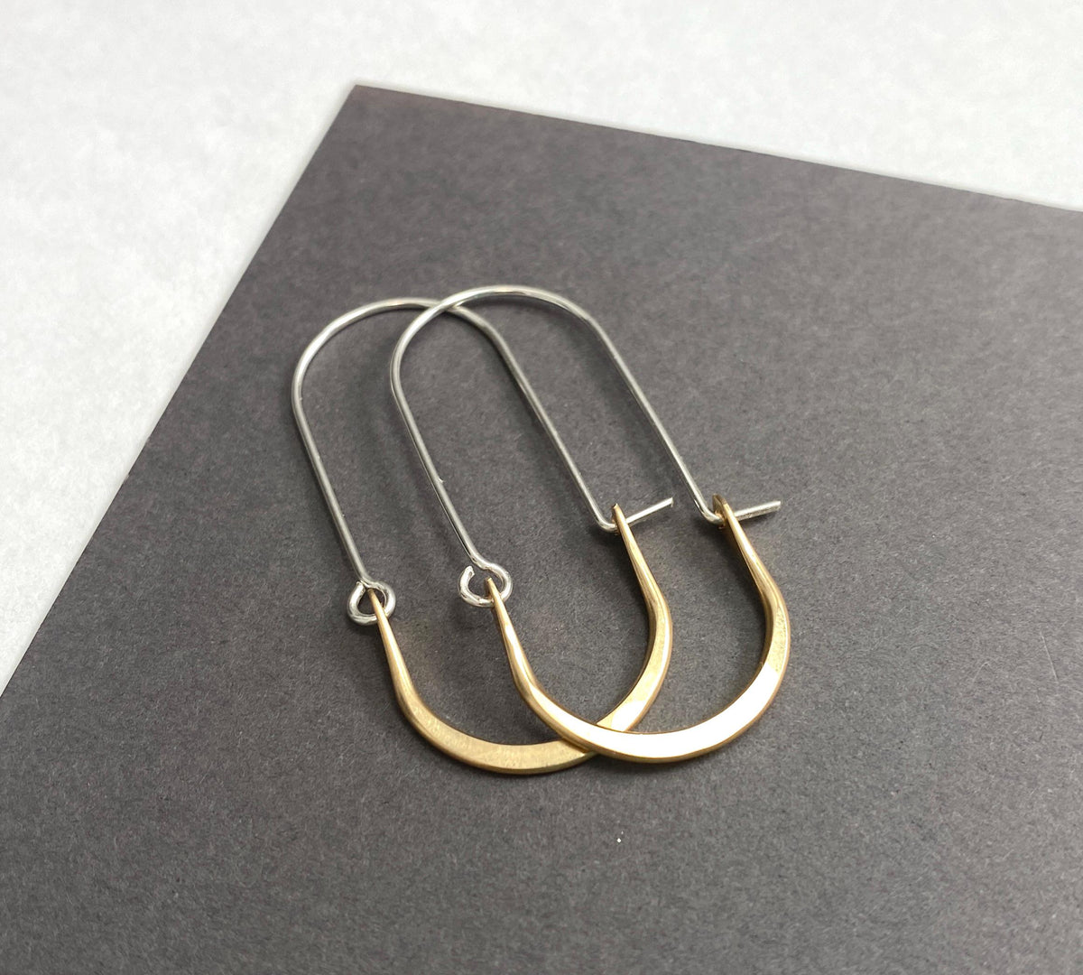 A pair of minimalist U-shaped drop hoop earrings in gold and silver arranged on a dark gray textured card against a light gray background.